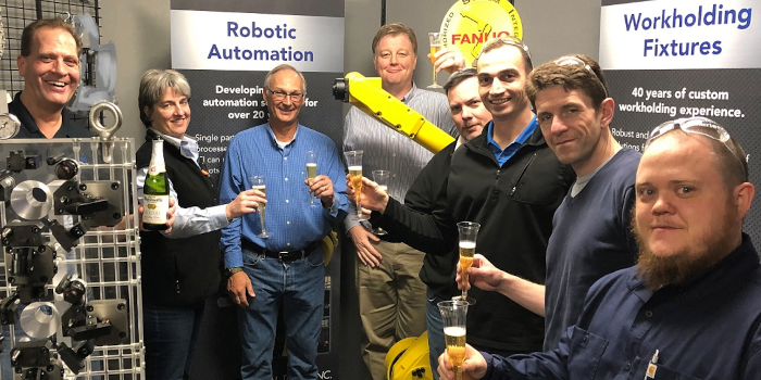A group of individuals in front of machinery, some holding glasses or bottles, indicating a celebratory gathering.